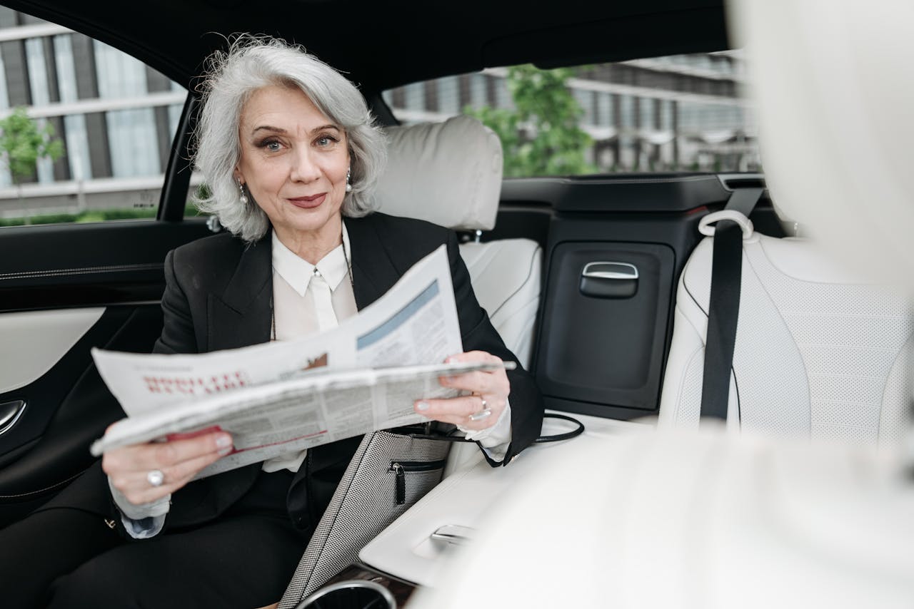 about-us Senior businesswoman reading a newspaper while seated in a car, smiling confidently.